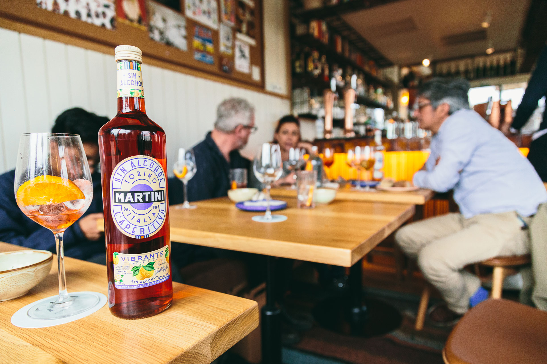 primer plano de botella de un aperitivo de martini sin alcohol y en el fondo se ven personas reunidas bebiendo en copas