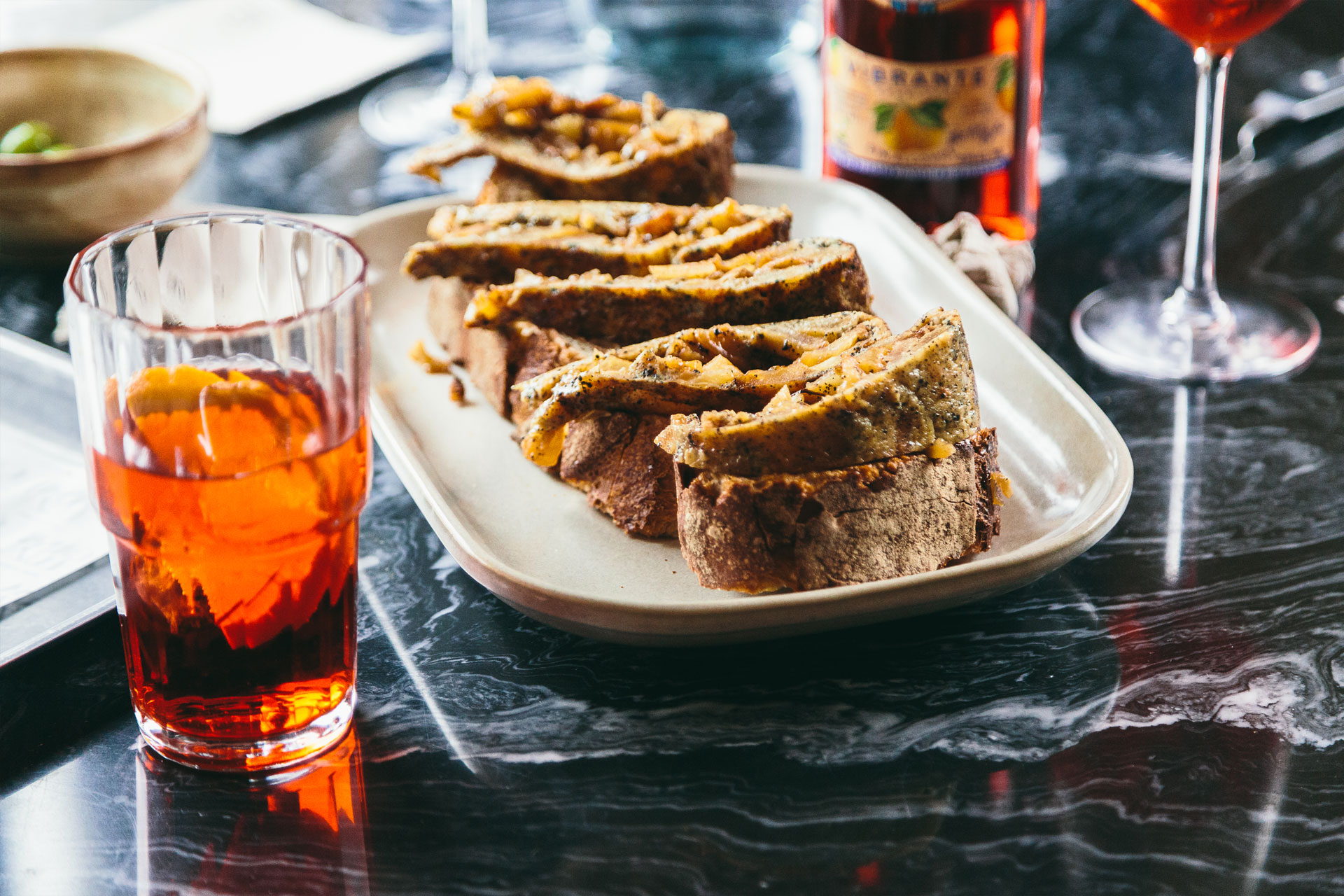 tapa de pan con tortilla sobre una mesa en la que se ve un vaso que lleva un aperitivo rojo de martini y una rodaja de limon