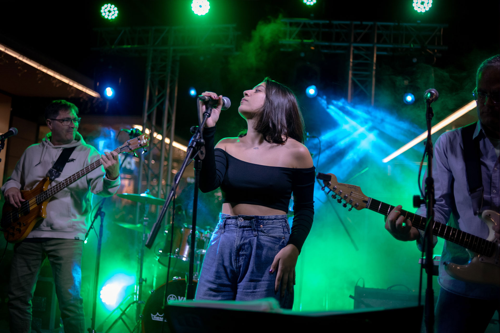 chica mira de lado mientras sujeta un microfono para cantar sobre un escenario situandose entre dos hombres tocando instrumentos
