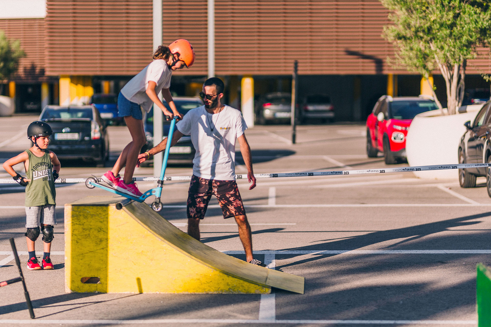 un parking en el que un niño mira a una niña que se tira de una rampa con patinete asistida de un chico joven que la ayuda a bajar